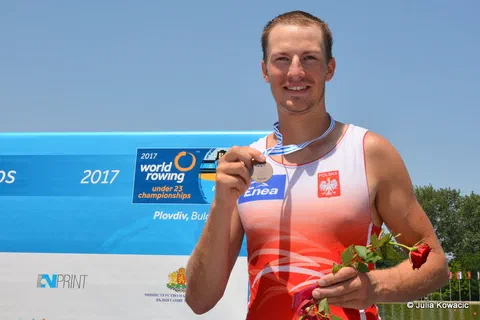 Uśmiechnięty wioślarz w biało-czerwonym stroju prezentujący medal na wstążce i czerwoną różę na podium
