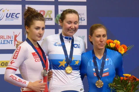 Trzy kolarki stojące na podium mistrzostw Europy z medalami na szyjach i bukietami kwiatów