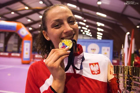 Uśmiechnięta zawodniczka w biało-czerwonej bluzie reprezentacji Polski całująca złoty medal obok pucharu